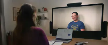 Two people in conversation using Starline. Person smiling on Starline display while engaged in conversation. The back of the head and hand of person in the foreground. There is a laptop and eyeglasses on the table between them.