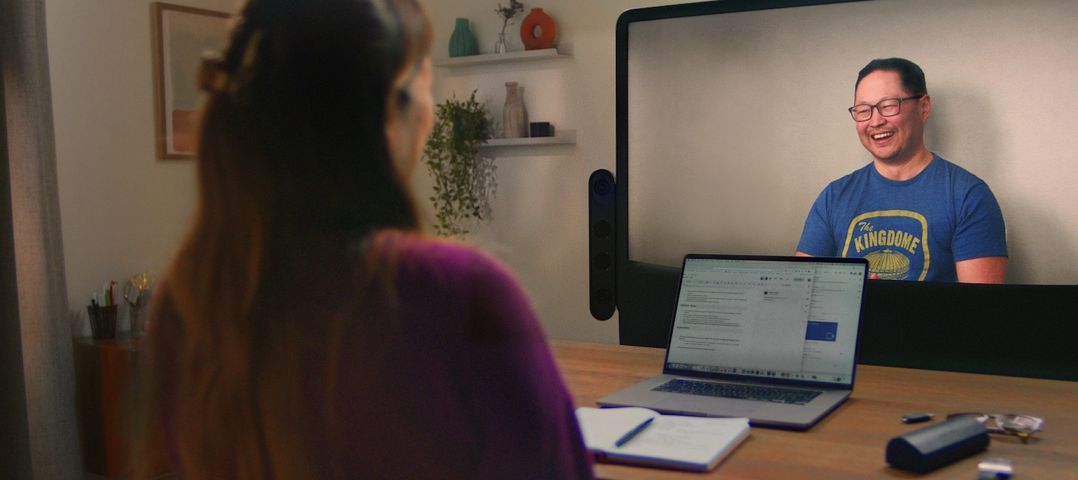 Two people in conversation using Starline. Person smiling on Starline display while engaged in conversation. The back of the head and hand of person in the foreground. There is a laptop and eyeglasses on the table between them.